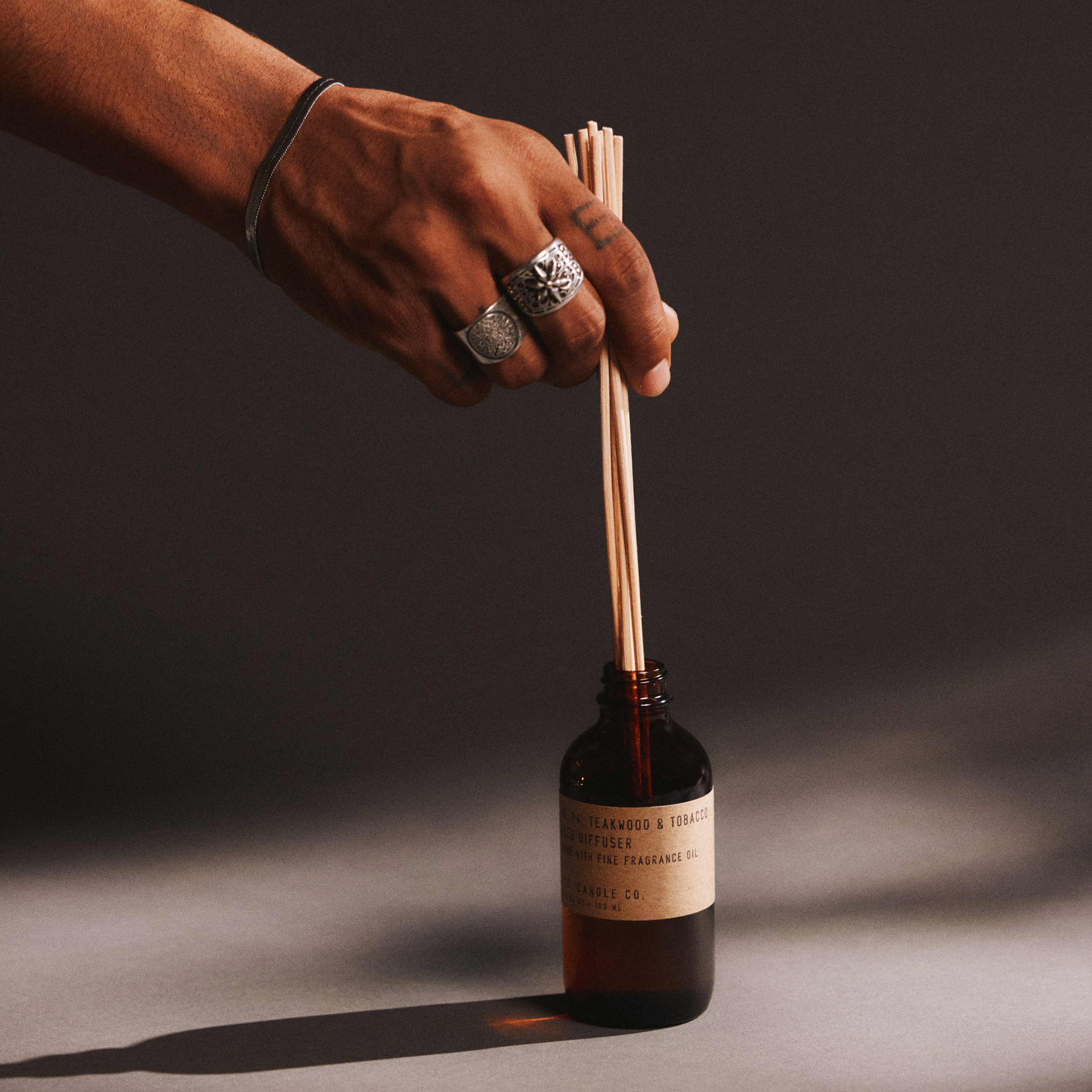 Hand with a ring holding reed diffuser sticks next to a bottle of teakwood & tobacco reed diffuser on a dark background